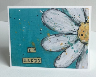May include: A turquoise blue background with white paint splatters. A white daisy with a yellow centre is painted on the right side of the card. The words "be happy" are written in black letters on a yellow background.