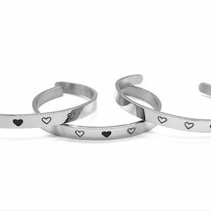 May include: Three silver-colored cuff bracelets with a row of black heart designs. The bracelets are open-ended and have a simple, minimalist style. The hearts are evenly spaced along the bands.