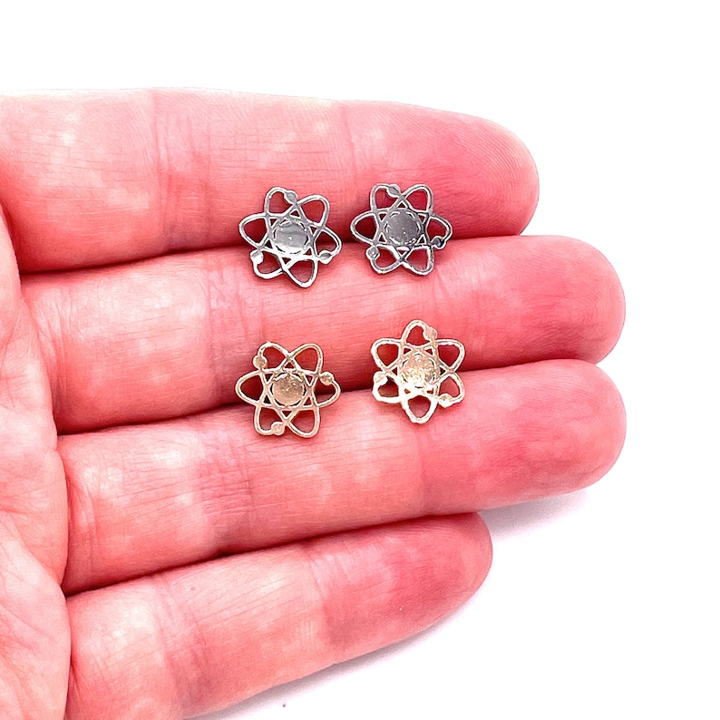 May include: A pair of earrings featuring a silver and gold atom design. The earrings are shown on a hand.