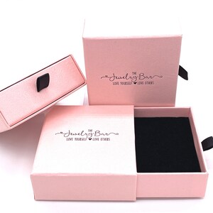 May include: Two pink jewelry boxes with a black velvet interior. The boxes have a drawer and a lid. The lid has the text "The Jewelry Bar Love Yourself Love Others" printed on it.