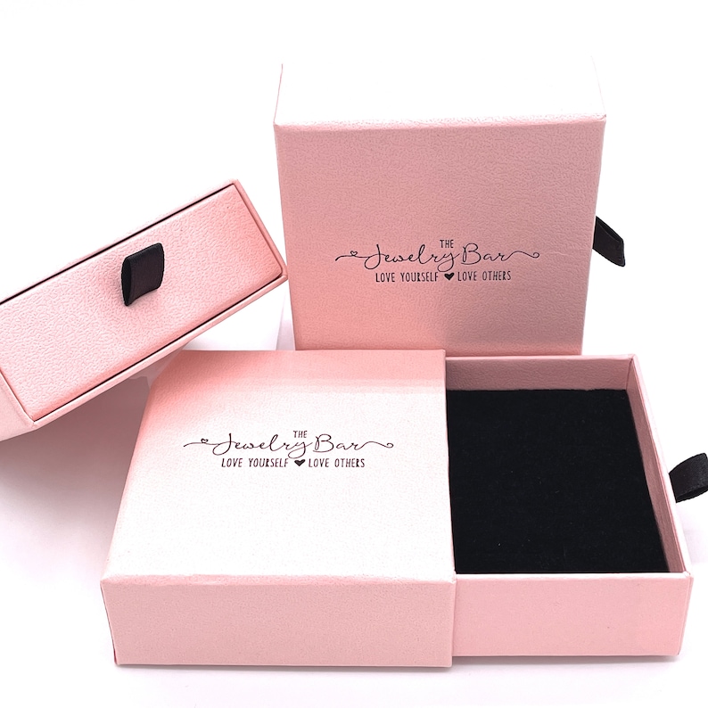 May include: Two pink jewelry boxes with a black velvet interior. The boxes have a drawer and a lid. The lid has the text "The Jewelry Bar Love Yourself Love Others" printed on it.