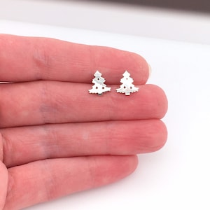 Puede incluir: Un par de pendientes de árbol de Navidad de plata. Los pendientes son pequeños y delicados, y presentan un diseño recortado de un árbol de Navidad.