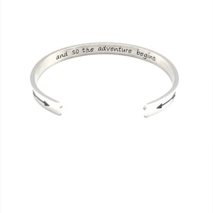 May include: Silver bangle bracelet with the inscription "and so the adventure begins" and two black arrows pointing in opposite directions.