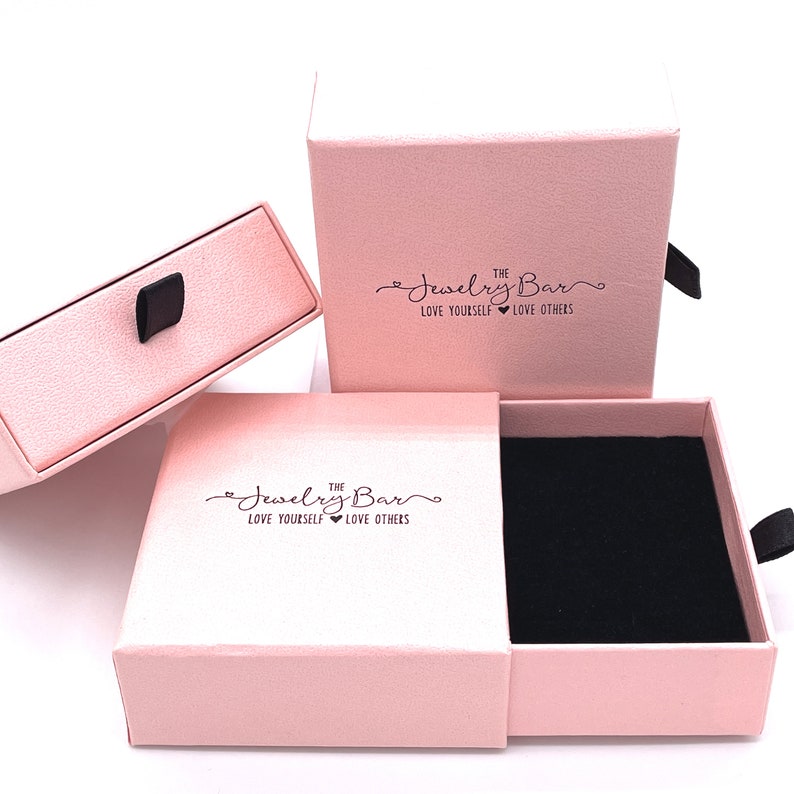 Puede incluir: Dos cajas de joyer&iacute;a rosas con un interior de terciopelo negro. Las cajas tienen un caj&oacute;n y una tapa. La tapa tiene el texto "The Jewelry Bar Love Yourself Love Others" impreso.