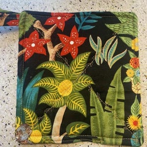 Puede incluir: Un posavasos acolchado negro con un estampado tropical con flores y hojas rojas, amarillas y verdes. El posavasos tiene un asa de bucle.