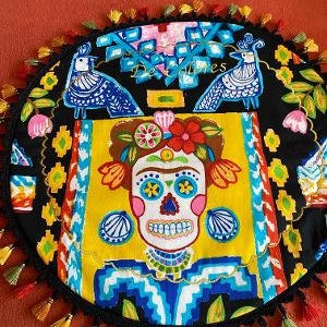 May include: A round black and yellow fabric cushion with a colorful design featuring a skull with flowers, birds, and geometric patterns. The cushion has a black fringe trim with colorful tassels.