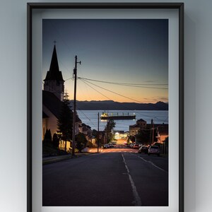 Peut inclure: Vue nocturne d'une rue dans une petite ville avec un clocher d'&eacute;glise en arri&egrave;re-plan. La rue est bord&eacute;e de maisons et d'arbres, et des voitures sont gar&eacute;es sur le c&ocirc;t&eacute; de la route. Un grand navire est visible au loin, naviguant sur l'eau.