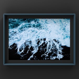 Peut inclure: Une photographie encadrée d'un gros plan de vagues blanches de l'océan qui s'écrasent sur des rochers noirs. L'image est prise d'un angle élevé, montrant l'eau blanche mousseuse et les rochers sombres et texturés en dessous.