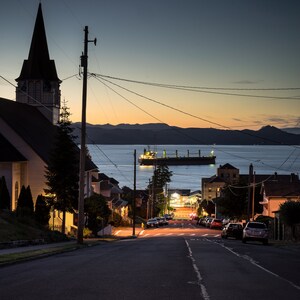 Peut inclure: Une silhouette sombre d'une &eacute;glise avec un clocher sur fond de ciel cr&eacute;pusculaire. Un grand navire de cargaison est amarr&eacute; au loin dans l'eau. Une rue bord&eacute;e de maisons descend vers l'eau.