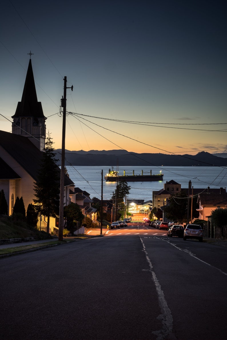 Peut inclure: Une silhouette sombre d'une &eacute;glise avec un clocher sur fond de ciel cr&eacute;pusculaire. Un grand navire de cargaison est amarr&eacute; au loin dans l'eau. Une rue bord&eacute;e de maisons descend vers l'eau.