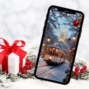 Può includere: Uno smartphone mostra una scena invernale con un tram su rotaie, alberi innevati e una cattedrale sullo sfondo. Una scatola regalo bianca con un nastro rosso e bacche rosse in primo piano.
