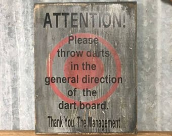 May include: A rustic wooden sign with a red circle and the text "ATTENTION! Please throw darts in the general direction of the dart board. Thank You, The Management."