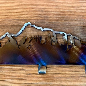 May include: Metal key rack featuring a mountain and trees silhouette design. The rack has five hooks and a dark blue, brown, and silver finish.