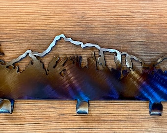 May include: Metal key rack with a mountain and trees silhouette design. The rack has five hooks and a dark blue, brown, and silver finish.