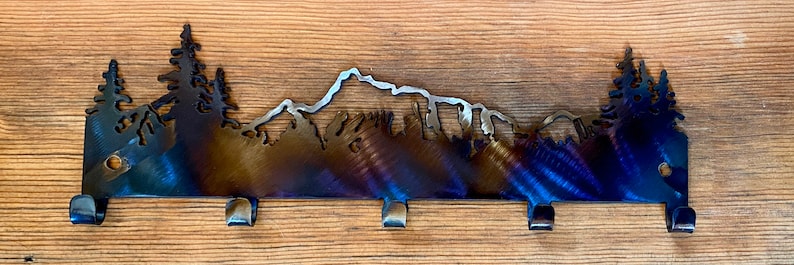 May include: Metal key rack featuring a mountain and trees silhouette design. The rack has five hooks and a dark blue, brown, and silver finish.