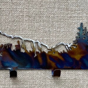 May include: Metal key hook featuring a silhouette of a mountain range and trees. The hook is dark blue with a silver finish. It has three hooks.