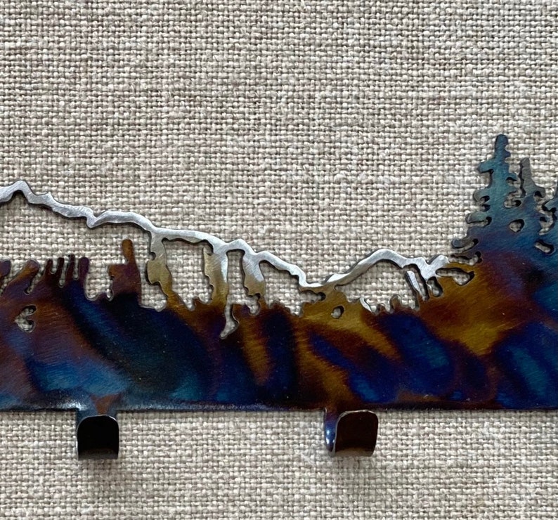 May include: Metal key hook featuring a silhouette of a mountain range and trees. The hook is dark blue with a silver finish. It has three hooks.