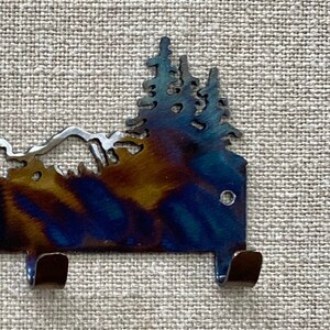 May include: Metal wall hook featuring a silhouette of a mountain range and pine trees. The hook is dark blue with a rainbow iridescent finish. It has two hooks on the bottom.