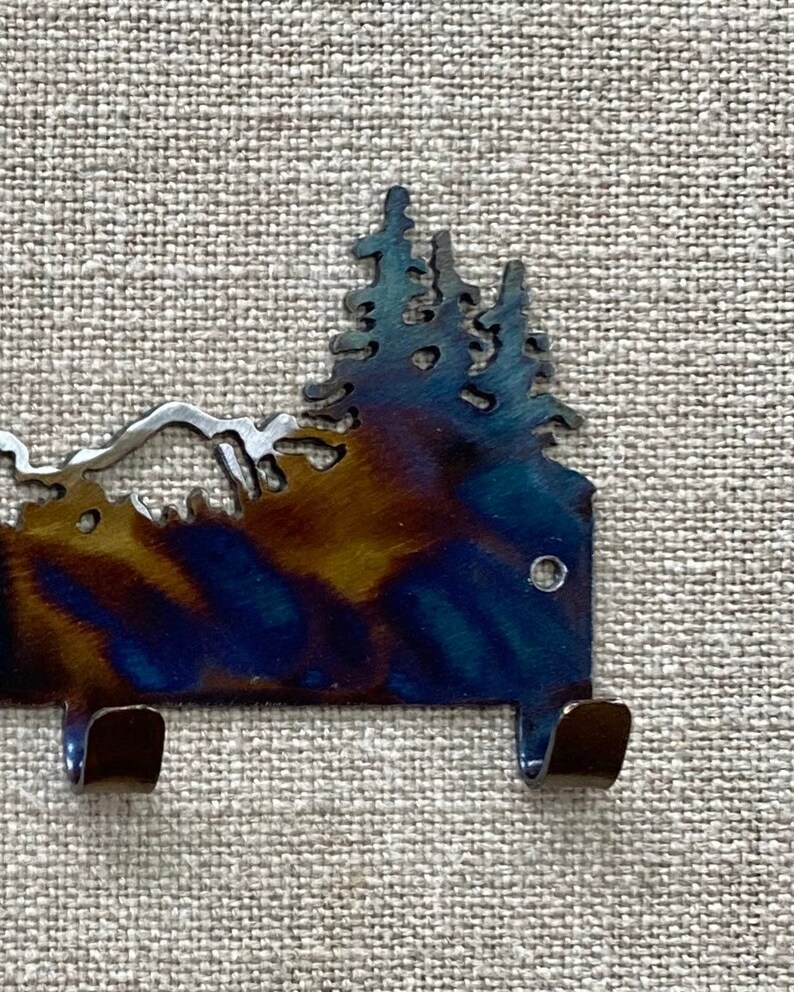 May include: Metal wall hook featuring a silhouette of a mountain range and pine trees. The hook is dark blue with a rainbow iridescent finish. It has two hooks on the bottom.