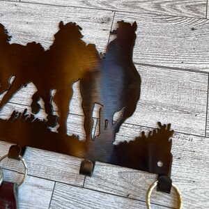 May include: Metal key holder with a silhouette of three horses. The key holder has four hooks for hanging keys. The key holder is painted in a dark brown color.