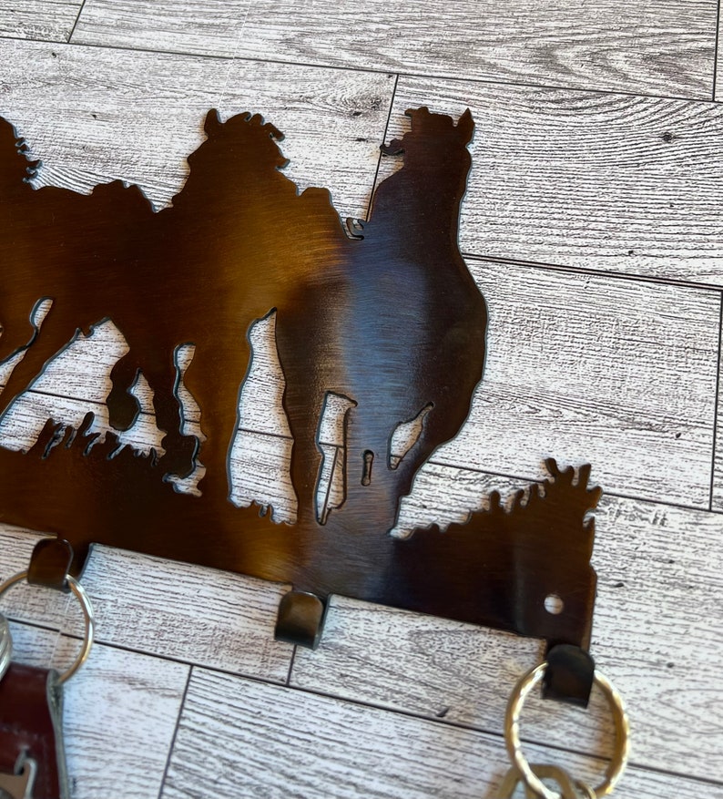 May include: Metal key holder with a silhouette of three horses. The key holder has four hooks for hanging keys. The key holder is painted in a dark brown color.