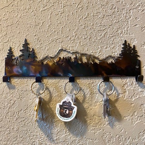 May include: A metal key rack shaped like a mountain range with trees and a bear silhouette. The rack has five hooks and is mounted on a wall. There are keys hanging on three of the hooks, including one with a Smokey Bear keychain that says "Only you can prevent forest fires."