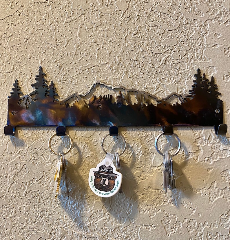 May include: A metal key rack shaped like a mountain range with trees and a bear silhouette. The rack has five hooks and is mounted on a wall. There are keys hanging on three of the hooks, including one with a Smokey Bear keychain that says "Only you can prevent forest fires."