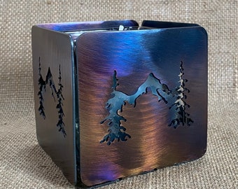 Pode incluir: Um porta-velas quadrado de metal com um design de montanha e pinheiros. O porta-velas tem um acabamento iridescente azul, roxo e marrom.