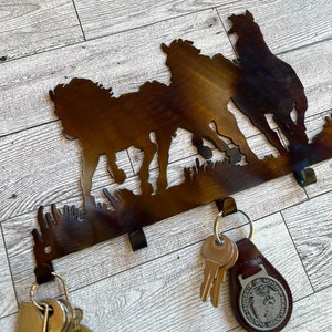 May include: A metal key holder with a silhouette of three horses running. The key holder has five hooks for hanging keys. The key holder is brown and has a rustic finish.