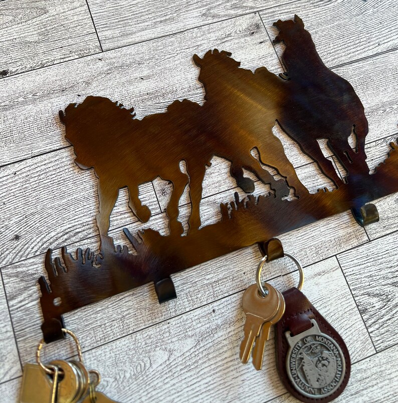 May include: A metal key holder with a silhouette of three horses running. The key holder has five hooks for hanging keys. The key holder is brown and has a rustic finish.