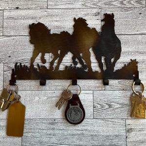 May include: Metal key holder with three horse silhouettes in a running pose. The holder has five hooks for hanging keys. There are keys and a key chain on the hooks.