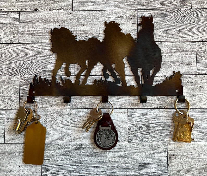 May include: Metal key holder with three horse silhouettes in a running pose. The holder has five hooks for hanging keys. There are keys and a key chain on the hooks.