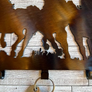 May include: Metal key holder with a silhouette of horses and a rustic brown finish. The key holder has four hooks and is mounted on a white wood background.