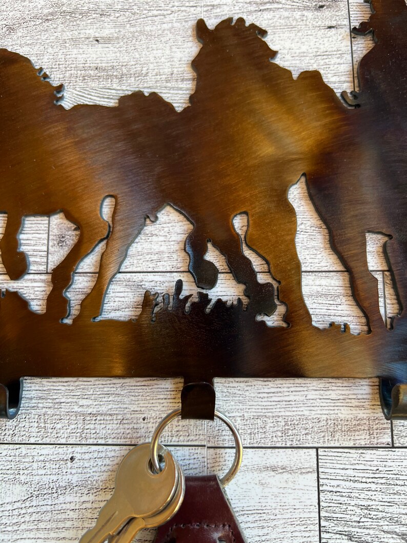 May include: Metal key holder with a silhouette of horses and a rustic brown finish. The key holder has four hooks and is mounted on a white wood background.