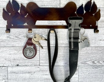 May include: A metal dog bone-shaped key holder with paw prints on either side. The holder has three hooks for hanging keys, leashes, or collars. A leash, keys, and a dog collar are hanging from the hooks.