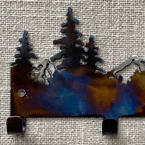 May include: Metal key rack featuring a mountain and pine tree design. The rack has a dark blue, brown, and gold finish and three hooks.