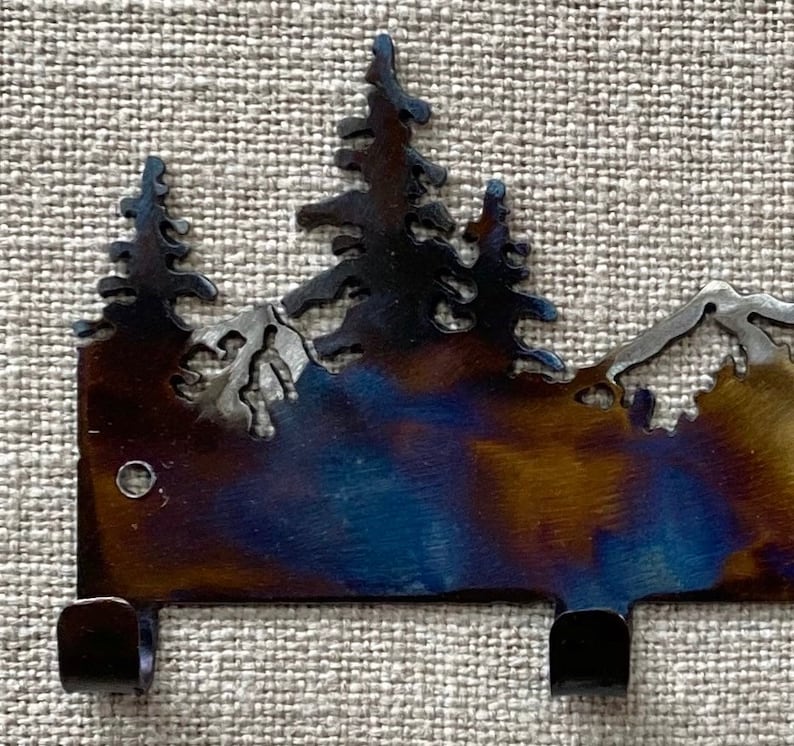 May include: Metal key rack featuring a mountain and pine tree design. The rack has a dark blue, brown, and gold finish and three hooks.