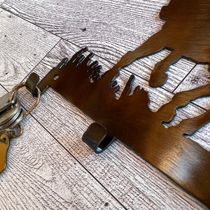 May include: A metal key holder shaped like a mountain range with a hook on the side. The key holder is brown and has a rustic finish. A set of keys is hanging on the hook.