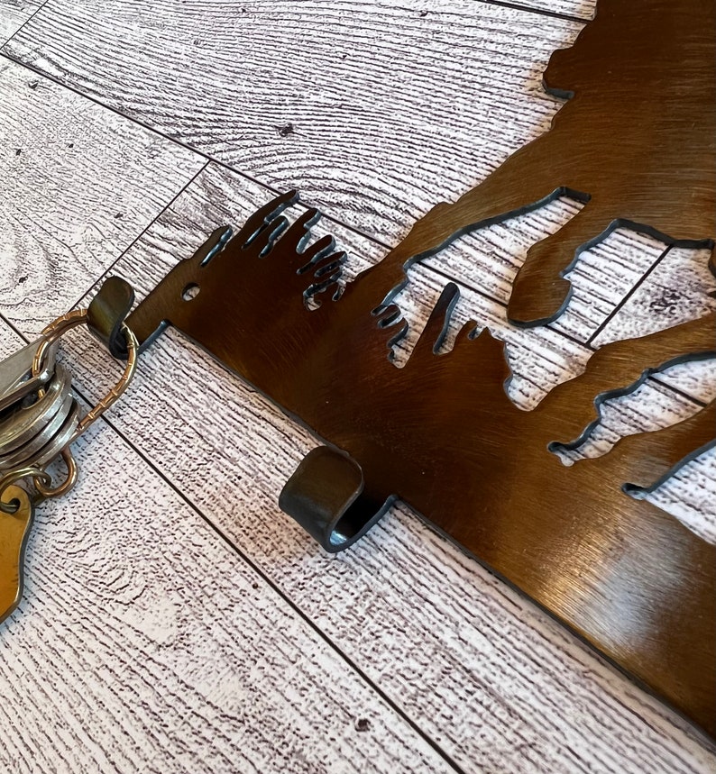 May include: A metal key holder shaped like a mountain range with a hook on the side. The key holder is brown and has a rustic finish. A set of keys is hanging on the hook.