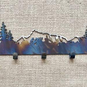 May include: Metal key rack featuring a mountain and tree silhouette design. The rack has five hooks and a rustic, burnt orange finish.