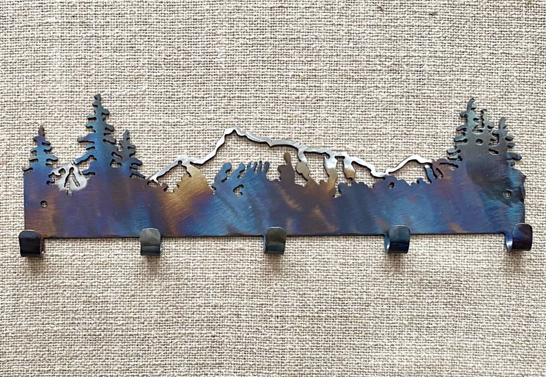 May include: Metal key rack featuring a mountain and tree silhouette design. The rack has five hooks and a rustic, burnt orange finish.