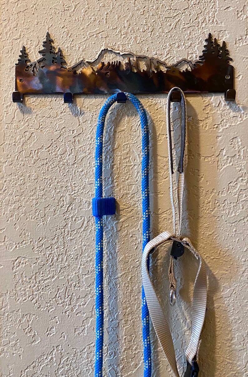 May include: A metal wall hook with a mountain range silhouette design. Two leashes, one blue and white rope and one beige nylon, are hanging from the hooks.