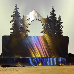 May include: Metal business card holder with a mountain and pine tree design. The holder has a brushed metal finish with iridescent rainbow hues.