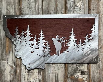 May include: Metal wall art featuring a silhouette of an elk standing in a forest, with a brown wood background. The elk is outlined in silver metal, and the trees are cut out of the metal to create a layered effect.