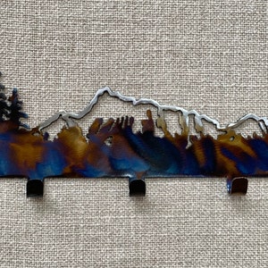 May include: A metal key rack featuring a mountain and tree silhouette design. The rack has a blue, brown, and silver finish and five hooks.