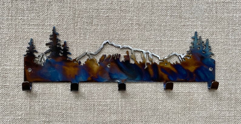 May include: A metal key rack featuring a mountain and tree silhouette design. The rack has a blue, brown, and silver finish and five hooks.