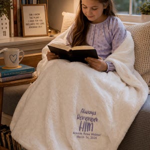 May include: A cozy, white, plush blanket with the embroidered text "Always Remember HIM" and a name and date. A young person is reading a book while wrapped in the blanket. A framed quote and a mug are visible in the background.