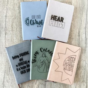 May include: A collection of five notebooks in various colors, including light blue, white, gray, green, and peach. Each notebook features embossed text and designs, such as "LOOK UNTO CHRIST," "HEAR HIM," and inspirational quotes. The notebooks have a textured appearance.