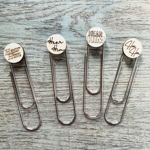 May include: Four silver-colored paper clips with wooden circular toppers. Each topper has the phrase "Hear Him" engraved in a different font. The clips are arranged on a light blue and gray wooden surface.