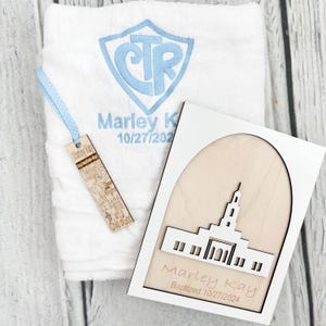 May include: A white towel with blue embroidered initials and name, a wooden bookmark, and a wooden plaque with a building design. The towel and plaque have the name "Marley Kay" and a date.